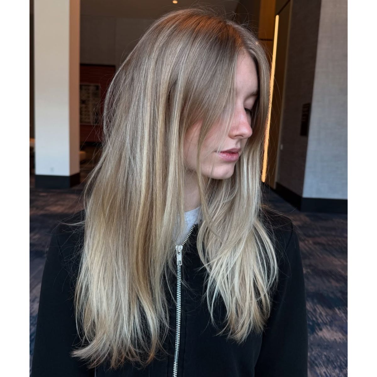 A woman with long, straight blonde hair featuring soft, natural highlights and a subtle root shadow, looking to the side in a softly lit indoor setting.