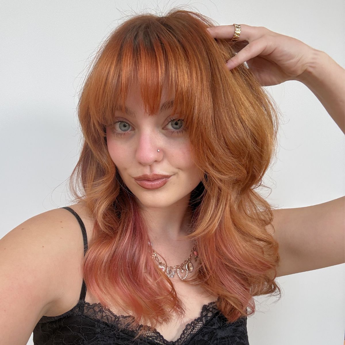 Model with long layered copper hair styled in soft waves with full fringe bangs and subtle peach-pink accents through the underneath sections.